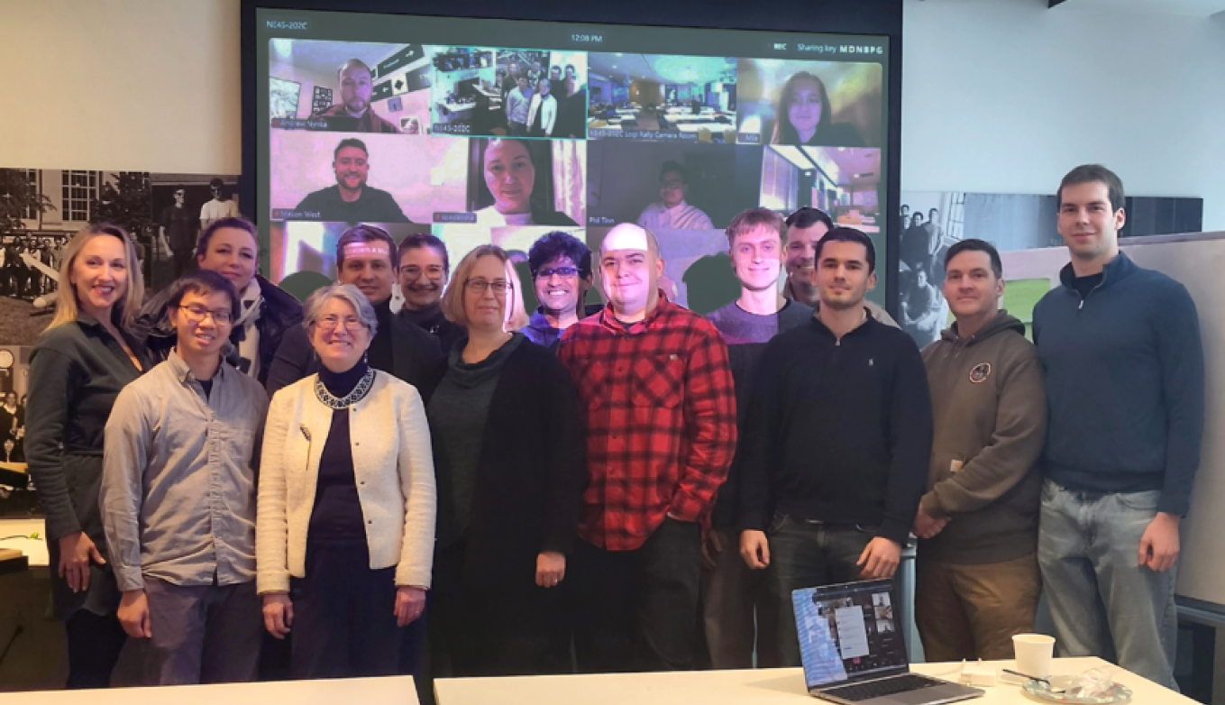 14 people stand around a large projection screen displaying at least a dozen other individuals on Zoom