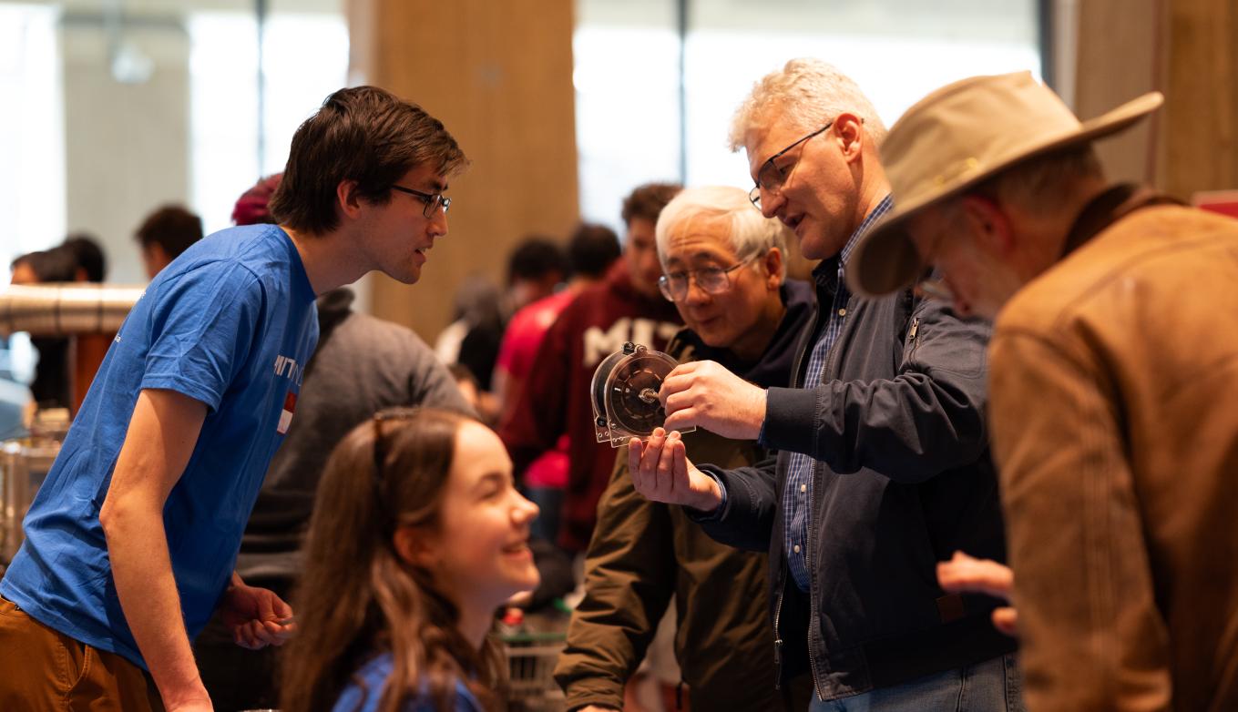 Members of the MIT Wind Team dressed in blue T-shirts chat with visitors, one of whom holds up a component part