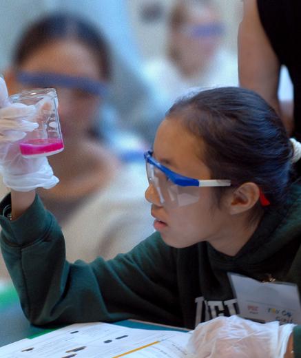 teenage girl in safety glasses and gloves examining the contents of a beaker