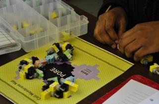 A pair of hands constructs proteins from colorful component pieces, arranged on top of a labeled mat