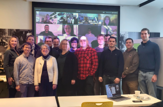 14 people stand around a large projection screen displaying at least a dozen other individuals on Zoom