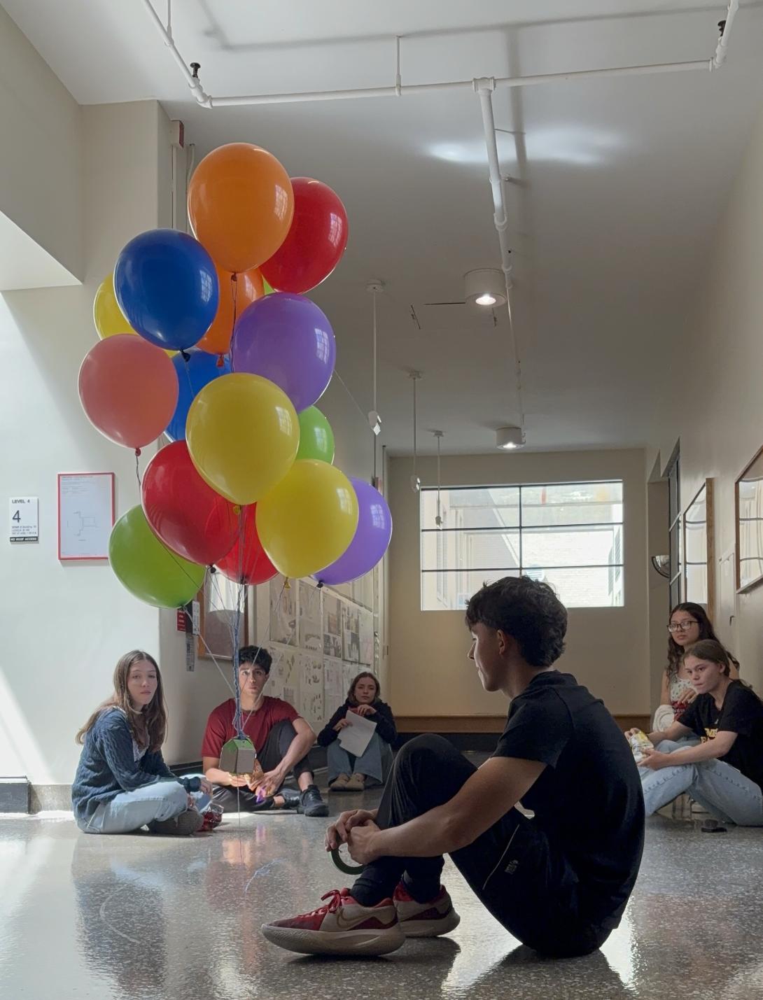 A group of students sit on the floor observing the buoyancy of a bunch of colorful balloons