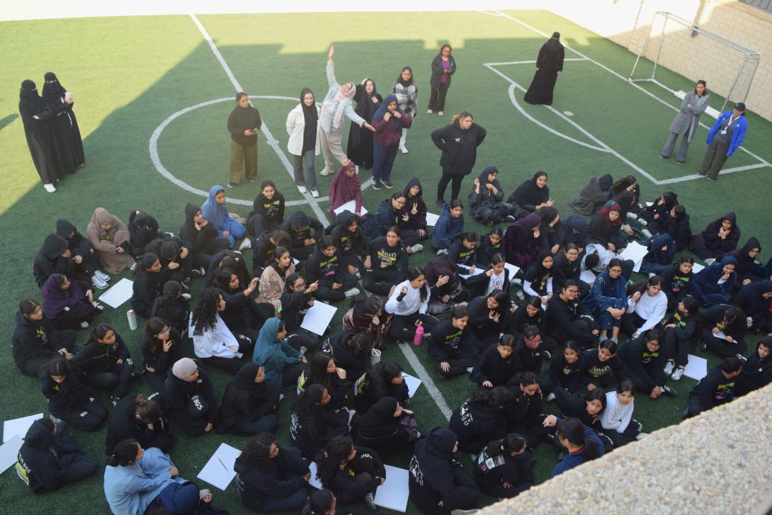 A group of students sits on a shaded soccer pitch in groups discussing hackathon ideas
